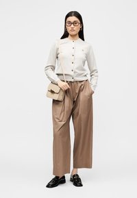 Mujer con cárdigan beige abotonado, pantalones anchos marrones, mocasines negros, gafas y lleva un bolso bandolera beige con decoración de animales.