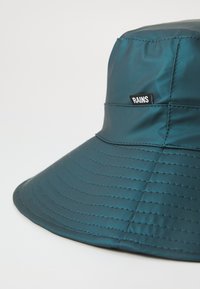 Türkiser Bucket Hat aus glänzendem Material, mit genähten Linien und einem kleinen schwarzen "RAINS"-Label an der Seite. Weiche Textur, breiter Rand.