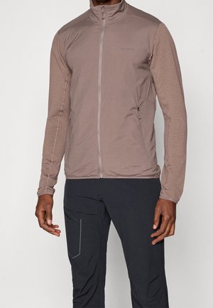 Homme portant une veste zippée marron clair avec des manches texturées et un pantalon noir avec une poche latérale et des détails réfléchissants.