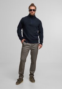 Navy blue knitted sweater with a high collar and half-zip, paired with light brown plaid trousers and green moccasin shoes.