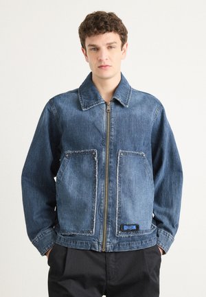Denim jacket, light blue, with a zip front, oversized fit, two front pockets, distressed edges, and a blue logo patch at the hem.