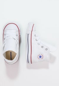 Pár bílých kotníkových tenisek Converse s červenými a modrými pruhy a viditelným logem značky, vystavených na světlém pozadí.