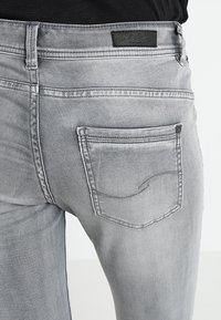 Jeans de mezclilla gris claro que presentan un bolsillo trasero con un patrón de costura curva y una etiqueta negra en la cintura, ofreciendo un diseño elegante y minimalista.