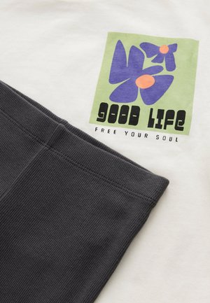 Camiseta de algodón en color crema con un cuadrado verde que presenta flores moradas y texto negro. Detalles de canalé en gris oscuro en el dobladillo.