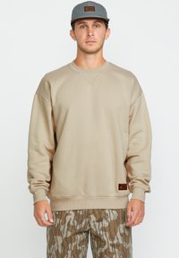 Hombre con sudadera beige, gorra gris con parche naranja y pantalones de camuflaje marrón de pie contra un fondo blanco.