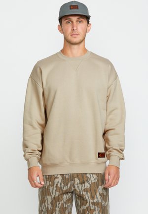 Mężczyzna ubrany w beżowy sweatshirt, szary kapelusz z pomarańczową łatą i brązowe spodnie w kamuflaż stoi na tle białego tła.