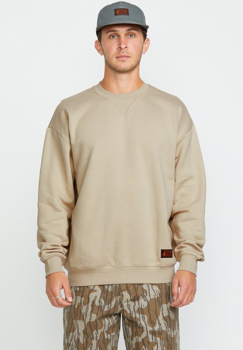 Hombre con sudadera beige, gorra gris con parche naranja y pantalones de camuflaje marrón de pie contra un fondo blanco.