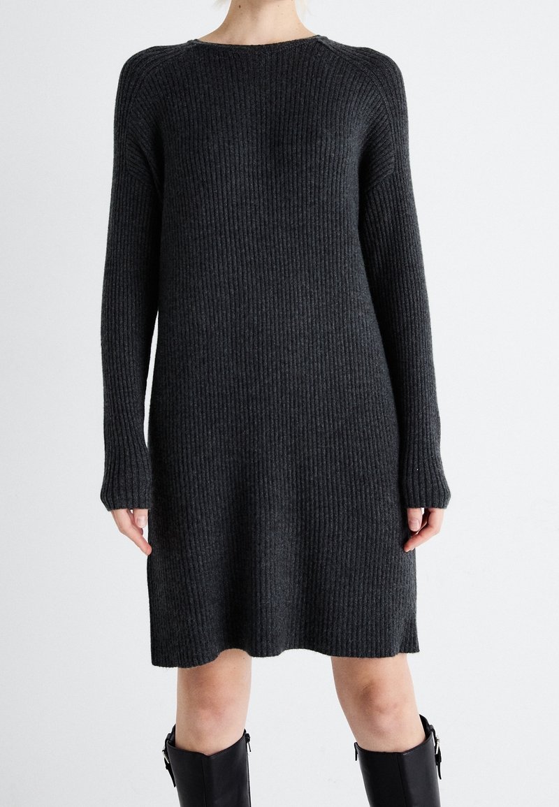 Femme portant une robe en maille côtelée gris foncé à manches longues, associée à des bottes noires montantes, se tenant devant un fond clair uni.