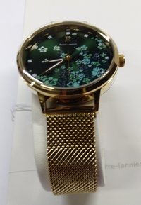 Reloj con correa de malla dorada y esfera de color verde oscuro, con patrones florales y agujas de hora y minuto doradas. Logo de la marca a las 12 en punto.