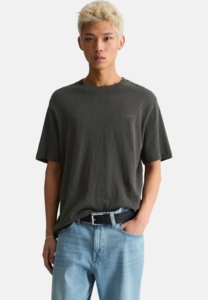 Giovane uomo con capelli biondi corti, indossa una t-shirt oversize grigio scuro e jeans azzurri con cintura nera, in piedi con una mano in tasca.