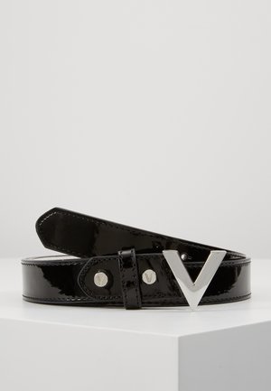 Musta kiiltävä nahkavyö hopeanvärisellä V-kirjaimen muotoisella soljella ja vastaavilla niiteillä, kierrettynä valkoisella pinnalla neutraalia taustaa vasten.