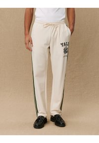 Cream jogger pants with green side stripes, featuring an elastic waistband with drawstring and a printed "Yale" logo on the front.