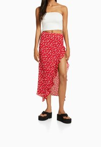 Punainen kukkakuvioinen kietaisuhame, jossa on röyhelöreuna ja sivuhalkio, yhdistettynä valkoiseen olkaimettomaan toppiin ja mustiin paksupohjaisiin sandaaleihin.