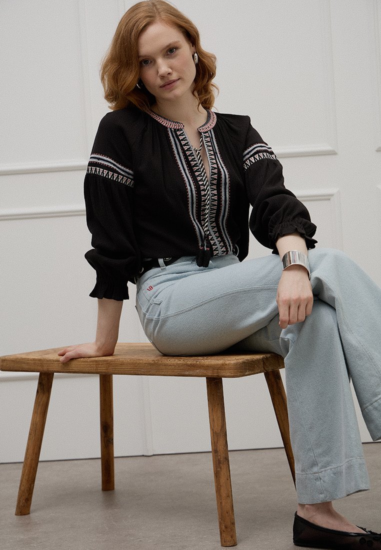 Schwarze Bluse mit gemusterter Stickerei, gerafften Ärmeln und rundem Ausschnitt, kombiniert mit hellblauen hoch taillierten Hosen. Hintergrund mit einem Holzstuhl.