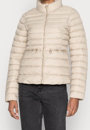 Beżowa pikowana kurtka z wysokim kołnierzem i przednim zamkiem błyskawicznym, z dwoma zamykanymi na zamek kieszeniami na wysokości talii, noszona z ciemnymi dżinsami.