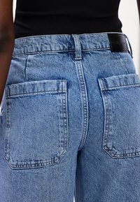 Jeans de mezclilla azul claro con cintura alta, grandes bolsillos traseros y un parche de cuero en la cintura. La textura es ligeramente desgastada con costuras visibles.
