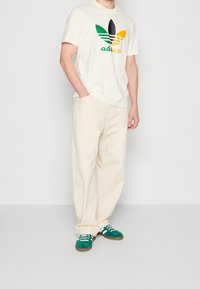Bílé tričko s vícebarevným logem Adidas, béžové kalhoty volného střihu a zelené tenisky s bílými tkaničkami a hnědou gumovou podrážkou.