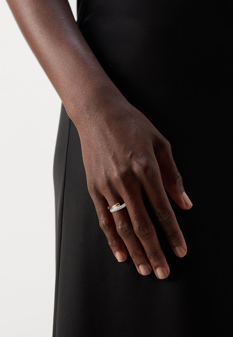 Bague en or et en argent présentant un bandeau central orné de diamants, posée sur une main reposant sur une surface en tissu noir. Texture lisse ; finition polie.