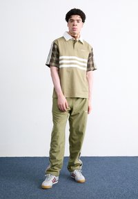 Olivgrünes Polohemd mit weißem Kragen, gestreiftem Vorderteil und karierten Ärmeln. Kombiniert mit olivgrünen Hosen und grauen Sneakern mit roten Akzenten.