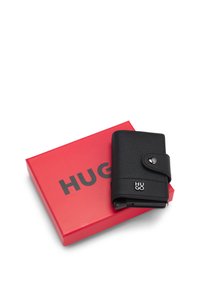 Billetera de cuero negro texturizado con cierre a presión y logo plateado de Hugo descansando sobre una caja roja con el texto "HUGO" en negrita y color negro.