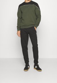 Olivgrön sweatshirt med svarta accenter och logotyp, tillsammans med svarta sweatpants med en logotyp på sidan, som bärs med svarta sneakers.