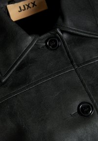 Veste en cuir noir au toucher lisse, dotée d'un col pointu et de deux boutons noirs à l'avant. Étiquette de marque visible à l'intérieur.