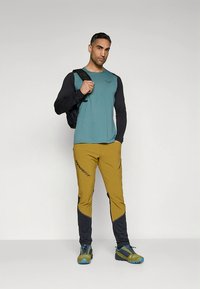 Man staat met een zwarte rugzak over één schouder, draagt een tealkleurig shirt met lange mouwen, gele en zwarte broek en blauwgroene trail schoenen.