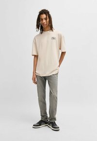 Beige t-shirt met korte mouwen en een zwart logo-opdruk op de linkerkant van de borst, gecombineerd met vervaagde grijze jeans en zwarte sneakers.