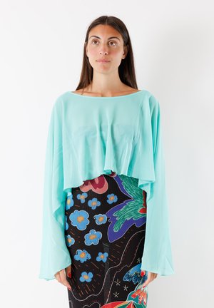 Donna dai lunghi capelli castani indossa una maglia azzurra a maniche lunghe e morbida e una gonna nera con fantasie floreali e astratte colorate.