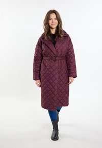 Palton burgundy matlasat cu talie cu centură, guler mare și mâneci lungi, purtat cu blugi albaștri și cizme negre până la genunchi.