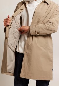 Beżowy płaszcz trencz z zapięciem na guziki, rozpięty, odsłaniający pikowaną szarą podszewkę i boczne kieszenie, noszony na białej bluzie z kapturem i ciemnych spodniach.