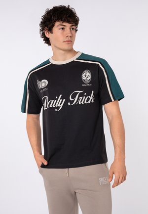 Junger Mann mit lockigem Haar, der ein schwarzes und grünes T-Shirt mit dem „Daily Trick“-Logo und beige Hosen trägt, die Hand in der Tasche hat und nach links schaut.