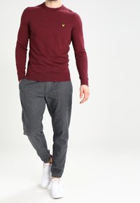Burgunderfarbene Strickjacke mit Rundhalsausschnitt, ausgestattet mit einem kleinen gelben Logo auf der Brust; kombiniert mit grauen strukturierten Hosen und weißen Sneakers.