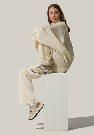 Top e pantaloni oversize di colore crema, abbinati a sneaker beige e nere con un design texturizzato e un marchio prominente sulla suola.