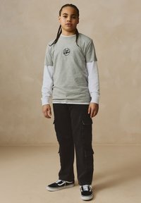 Gray short-sleeve t-shirt with black logo, layered over a white long-sleeve shirt. Black cargo pants and black sneakers with white soles.