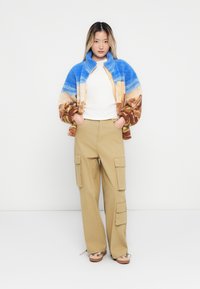 Jeune femme avec les mains dans les poches, portant une veste en polaire bleue et marron, une chemise blanche, un pantalon cargo beige et des chaussures à motifs sur fond blanc.