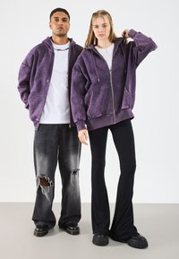 Felpa viola oversize con zip, tasche frontali e cappuccio con cordino; abbinata a pantaloni neri aderenti e jeans grigi strappati.