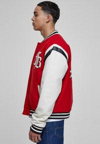 Rød og hvid varsityjakke med ribbede manchetter. Har forknapper, broderet logo og sorte stribede detaljer på ærmerne.