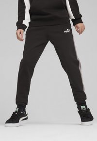 Pantalons de jogging noirs avec un logo Puma blanc, une taille élastique et des rayures latérales grises. La matière semble douce et texturée.