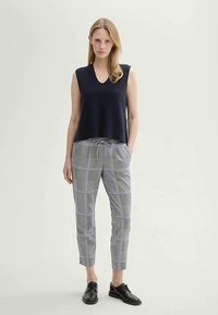 Ärmlös marinblå stickad topp med V-ringning, kombinerad med grå rutiga skräddarsydda byxor med dragsko i midjan och svarta snörskor.