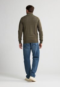 Dunkelolivgrüner Strickpullover mit hohem Kragen, kombiniert mit hellblauen Straight-Leg-Jeans und beigen Sneakers, von hinten betrachtet.