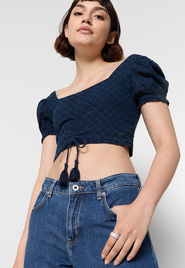 Marineblaues Cropped Top mit Puffärmeln, verziert mit einer Augenmustertextur und einem Krawattenakzent mit Quasten. Kombiniert mit hochtaillierten Denim-Jeans.