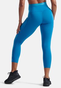 Blå högmidjade leggings med en mjuk textur och cropped längd. Har en logotryck vid vristen och kombineras med svarta träningsskor.