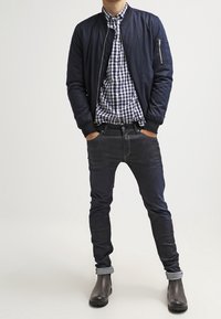 Giacca bomber navy sopra una camicia a quadri, jeans scuri e stivaletti neri. La giacca ha una tasca con zip; i jeans hanno i polsini arrotolati.