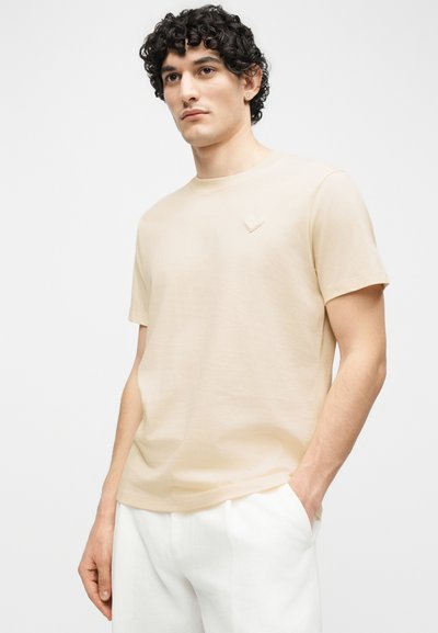 Άνδρας με σγουρά σκούρα μαλλιά που φοράει μπεζ κοντομάνικη μπλούζα και λευκό παντελόνι, στέκεται με το ένα χέρι στην τσέπη.