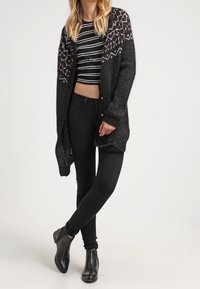 Cárdigan negro de punto con un diseño en la parte superior, combinado con una camiseta de cropped a rayas blancas y negras y jeans negros ajustados. Botines negros completan el look.