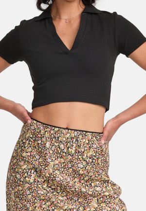 Kobieta ubrana w czarną, krótko skrojoną koszulkę polo oraz wysokogównową kwiecistą spódnicę w różowe, żółte i zielone kwiaty na jasnym tle.