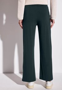 Pantaloni a gamba larga in tessuto verde scuro, con una texture liscia e una vita elasticizzata. Indossati con sneakers bianche, mostrati da dietro.