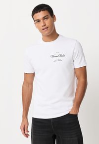 Fehér rövid ujjú pamut póló, középen a mellkasnál fekete nyomott logóval, sötét farmernadrággal párosítva.