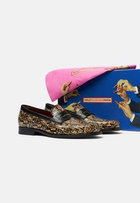 Mocasines negros con un patrón dorado de instrumentos musicales, con un diseño de doble hebilla. Acompañados de un embalaje colorido que muestra claramente manos.